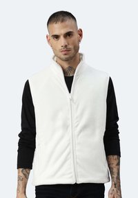 Weißer Fleece-Weste mit hohem Kragen, Reißverschluss und seitlichen Taschen, getragen über einem schwarzen Langarmshirt. Verfügt über eine glatte Textur.