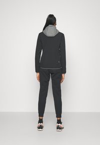 Veste noire avec capuche grise, fermeture éclair et poignets élastiques, associée à un pantalon de survêtement noir et des chaussures de sport multicolores.