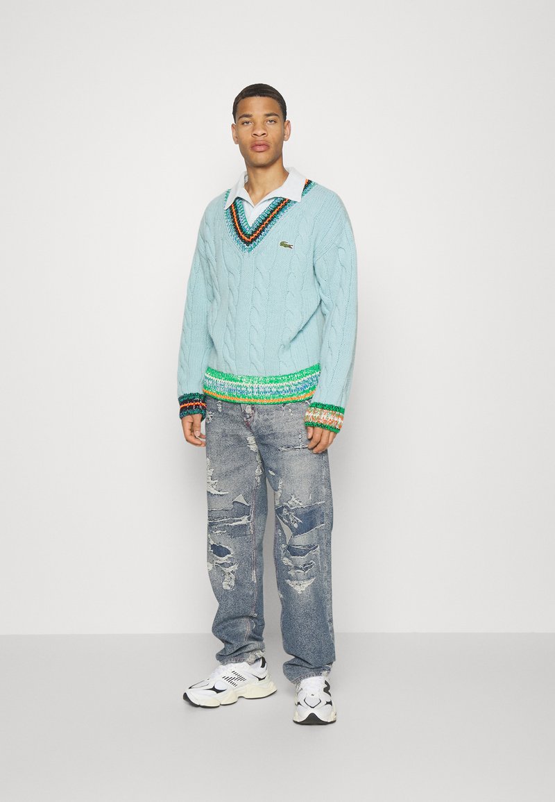 Uomo in piedi contro uno sfondo bianco, che indossa un maglione in cotone leggero azzurro a treccia con dettagli multicolori, jeans strappati e sneaker bianche.