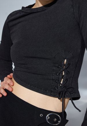 Top corto nero a maniche lunghe, presenta dettagli in pizzo laterali, realizzato in morbido tessuto di cotone, con scollatura rotonda e design aderente.