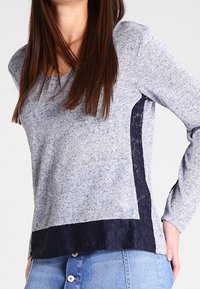 Grau langärmeliges Oberteil aus strukturiertem Stoff, mit einem dunkelblauen Spitzenbesatz an den Seiten und einem einfarbigen dunkelblauen Saum am unteren Rand.