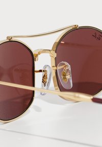 Slnečné okuliare Ray-Ban s zlatým rámom a tmavohnedými sklami, s nastaviteľnými priehľadnými nosovými podložkami na bielom povrchu.