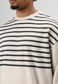 Sweat-shirt beige avec des rayures noires horizontales, col rond et texture douce. Présente une coupe décontractée et des épaules tombantes.