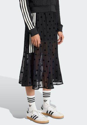 Personne portant une jupe midi transparente noire à pois, une veste noire avec des rayures blanches sur les manches, des chaussettes blanches Adidas et des baskets blanches.