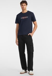 Tamno plava pamučna majica s bijelim i crvenim Guess Jeans logotipom; uparen s crnim cargo hlačama sa bočnim džepovima. Bijele tenisice s smeškastim naglascima.