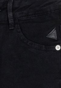 Svarta denimjeans med en framficka som har en triangulär logotypapplikation och en synlig metallknapp som accent. Slät textur och figurnära design.