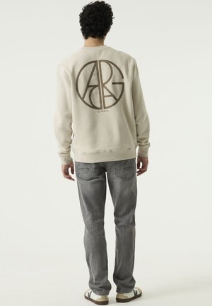 Man with curly hair stands facing away, wearing a beige sweatshirt with a large circular logo, gray jeans, and white sneakers.