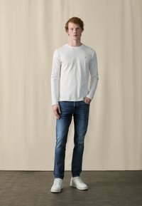 Hvid langærmet skjorte, blå jeans og hvide sneakers. Enkel design med rund halsudskæring og lille logo på brystet. Ren, afslappet look.