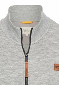 Veste zippée grise avec un col haut, tissu texturé, zip noir avec tirette en cuir, et une touche orange à l'intérieur du col.