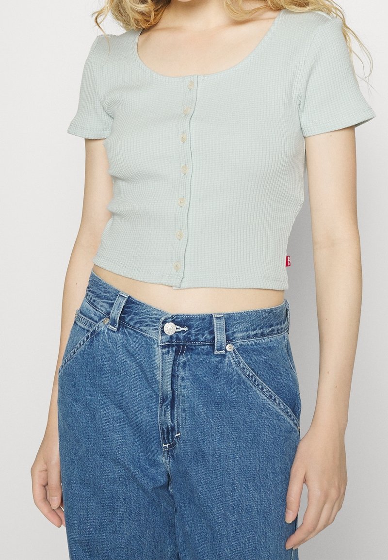 Světle modrý, žebrovaný cropped top s výstřihem typu scoop a krátkými rukávy, který má zapínání na knoflíky vpředu a štítek na dolní straně.