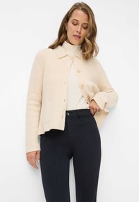 Beige gebreid vest met een kraag en knopen aan de voorkant. Heeft lange mouwen, een zak en een korte zoom, gecombineerd met zwarte broeken.