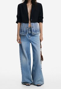 Jeansjacke mit einem schwarzen, kurz geschnittenen Oberteil und einem langen, blauen Jeansstoff unten. Weit geschnittene blaue Jeans mit ausgewaschener Textur. Sandalen mit dicken Absätzen.