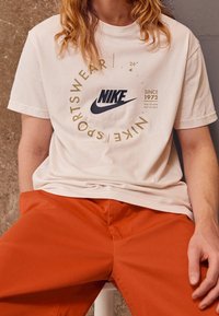 T-shirt en coton blanc présentant un logo Nike bleu foncé et un texte doré. Le modèle porte un pantalon orange. L'arrière-plan a un mur texturé gris.