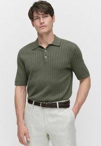 Olivgrön ribbad pikétröja med korta ärmar, treknappad knäppning, bärs med ljusa linbyxor och ett flätat brunt bälte.