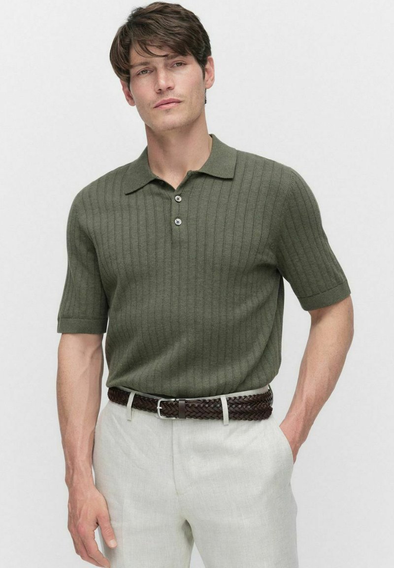 Olivgrön ribbad pikétröja med korta ärmar, treknappad knäppning, bärs med ljusa linbyxor och ett flätat brunt bälte.