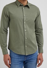 Man in een olijfgroene button-up langemouwen shirt met het 'Lee' logo op de borst, gecombineerd met een blauwe spijkerbroek.