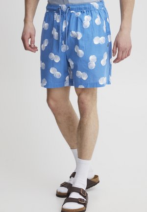 Blue swim shorts with white shell-pattern print, elastic waistband, and drawstring. Worn with white socks and brown sandals.