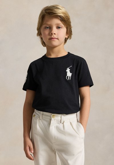 Blond barn iført en sort polo med en hvid hestelogo og hvide bukser, stående med den ene hånd i en lomme mod en ensfarvet baggrund.