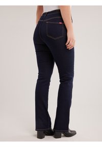 Osoba odjevena u tamnoplave uske traperice i crne čizme s potpeticom, stojeći s jednom rukom opuštenom uz tijelo na jednostavnoj pozadini.