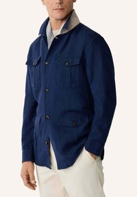 Mand iført marineblå knap-op jakke med fire knap-lukkede lommer, lyse bukser, hånd i lommen, beige krave synlig under jakkens krave.