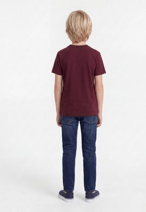 Barn med blondt hår vendt væk iført en mørkerød t-shirt, blå jeans og mørke sneakers på en hvid baggrund.