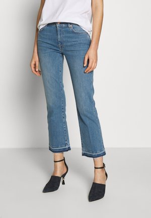 Vrouw draagt een blauwe cropped flare jeans met onafgewerkte zomen, een wit bovenstuk en zwarte enkellaarsjes met hak, staand tegen een effen muur.