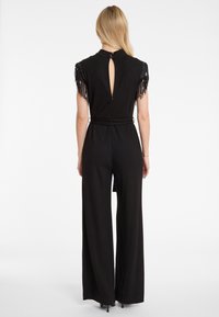 Zwart jumpsuit met franjes aan de mouwen, open rug en een ceintuur in de taille. Het wijdvallende ontwerp reikt tot de vloer, wat een vloeiende uitstraling geeft.