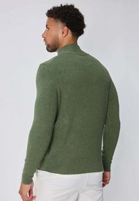 Homme avec des cheveux bouclés courts portant un pull en maille vert ajusté et un pantalon blanc, tourné sur le côté devant un fond uni.
