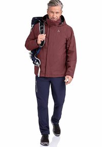 Weinrot wasserdichte Jacke mit hohem Kragen, kombiniert mit blauen Hosen und schwarzen Schuhen. Das Modell trägt einen blauen Rucksack.