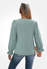Jasnozielona bluzka z teksturowanymi wzorami w diamenty i kwiaty, z bufiastymi długimi rękawami i falbanami na mankietach, w połączeniu z niebieskimi dżinsami i małą torebką.