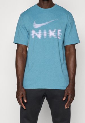 Homme portant un t-shirt Nike bleu avec un logo blanc style glitch et un pantalon noir, debout devant un fond uni.