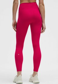 Prijete roza legice iz elastičnega materiala, z visokim pasom in majhnim logotipom blizu gležnja, v kombinaciji z lahkimi športnimi copati.