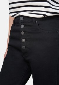 Jean noir à taille haute et fermeture à cinq boutons. Présente une texture lisse, deux poches avant et des détails de couture bien marqués.