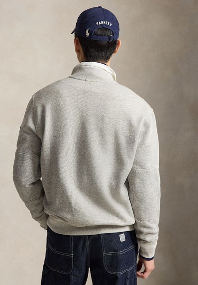 Homme portant un sweat-shirt gris clair et un jean bleu foncé, de dos, avec une casquette de baseball des Yankees bleu marine sur la tête.
