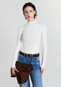 Frau, die ein weißes geripptes Langarmoberteil, blaue Jeans, einen braunen Gürtel trägt und eine braune Wildlederjacke vor einem schlichten hellen Hintergrund hält.