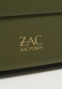 Olivově zelený kožený povrch s béžovým logem "ZAC ZAC POSEN" a viditelným stehem podél horního okraje.