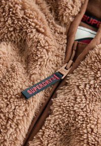 Beżowa polarowa kurtka z teksturowaną, puszystą powierzchnią. Posiada brązowy zamek i czarną metkę z napisem "SUPERDRY" w czerwonym kolorze.