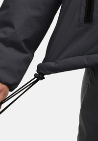 Giacca leggera nera con coulisse in fondo e tasca con zip sul petto. Il tessuto ha una texture liscia e opaca.