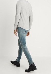 Camisa gris de manga larga, diseño ajustado. Vaqueros claros desgastados con un corte entallado, con detalles de salpicaduras de pintura. Botas negras hasta el tobillo.