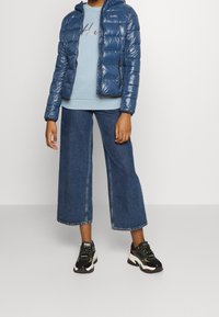 Blaues Puffer-Jackett mit glänzender Oberfläche und Kapuze, kombiniert mit einem hellblauen Sweatshirt, weit geschnittenen Jeans und schwarzen Sneakern mit grünen Akzenten.