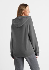 Grauer Kapuzenpullover mit lockerer Passform, gerippten Bündchen und Saum sowie weichem Material. Die Rückansicht zeigt ein simples Design und eine neutrale Farbe.