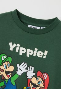 Groene katoenen t-shirt met cartoonfiguren Mario en Luigi, met de tekst "Yippie!" in vet, witte letters boven hen.