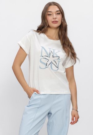 Jeune femme posant en chemise blanche à manches courtes avec motif étoile de mer et pantalon taille haute bleu clair sur fond uni.