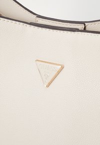 Borsa in finta pelle beige chiaro con texture, caratterizzata da un logo Guess triangolare color oro. Design semplice con cuciture pulite e accenti minimi.