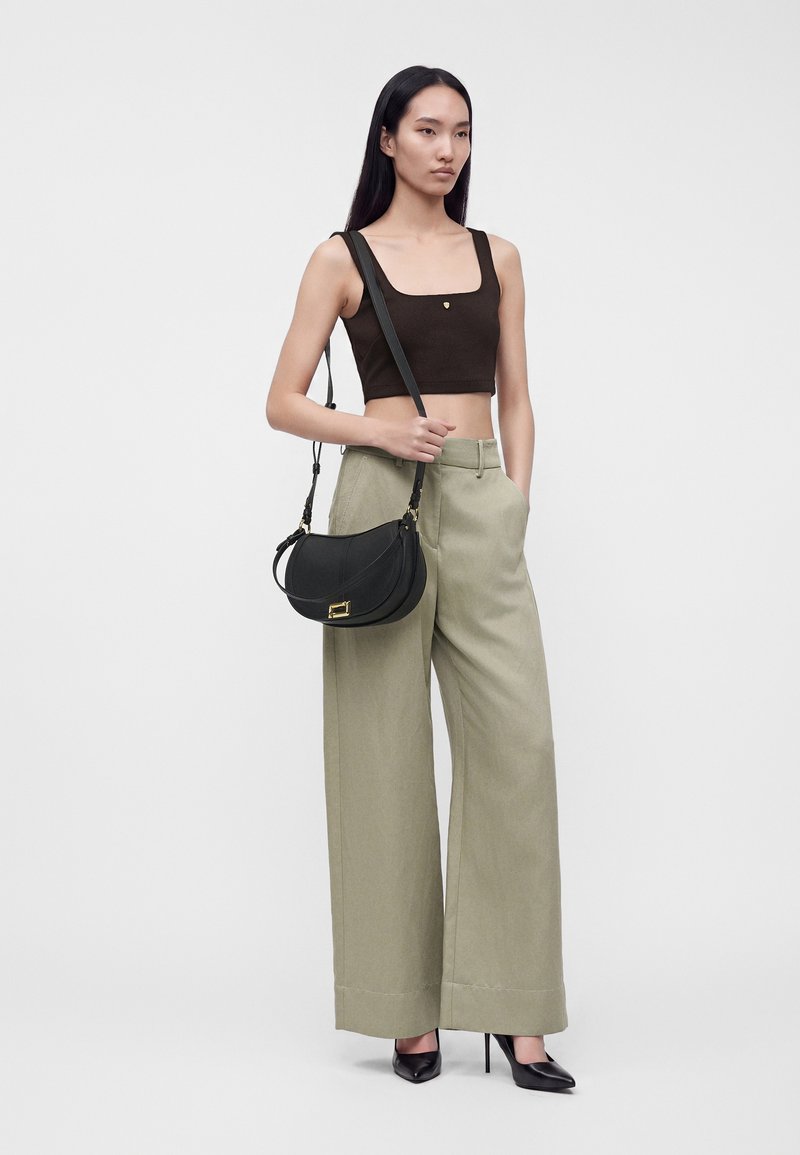 Femme portant un crop top noir, un pantalon large beige, des talons noirs et un sac à bandoulière noir avec des ferrures dorées.