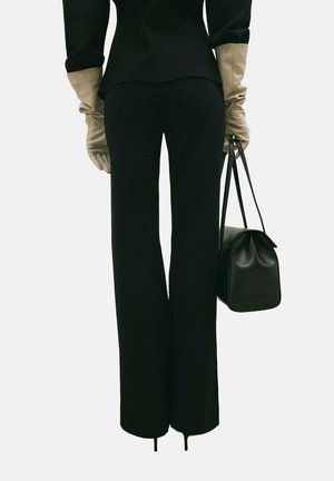 Chaqueta de traje negra ajustada en la cintura con mangas largas, combinada con pantalones negros acampanados. Accesorios incluyen guantes beige y un pequeño bolso negro.