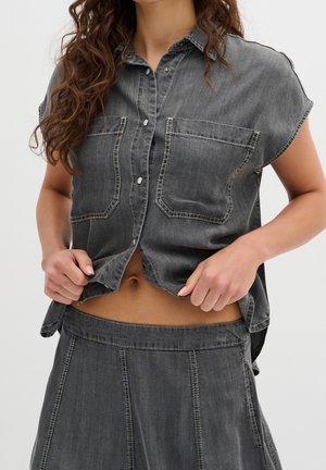 Kobieta ubrana w wyblakłą czarną koszulę z krótkim rękawem z denimu na guziki oraz pasującą, wysokowaisted spódnicę, trzymająca lekko uniesiony brzeg koszuli.