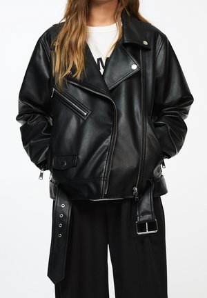 Personne portant une veste de motard en cuir noir avec des fermetures éclair et des boutons argentés, les mains dans les poches, par-dessus une chemise blanche et un pantalon noir.