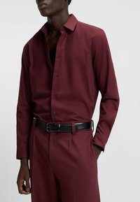 Burgundy long-sleeve shirt with a button-down collar, front buttons, and a black belt. Smooth texture with a tailored fit and side pockets.