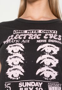 Svart bomull t-shirt med ett tryckt mönster i rosa, som visar flera ögonillustrationer och textdetaljer för ett evenemang.